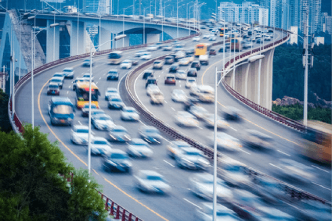 Verkehr auf einer geschwungenen Autobahn mit vielen Fahrzeugen und städtischer Umgebung im Hintergrund.