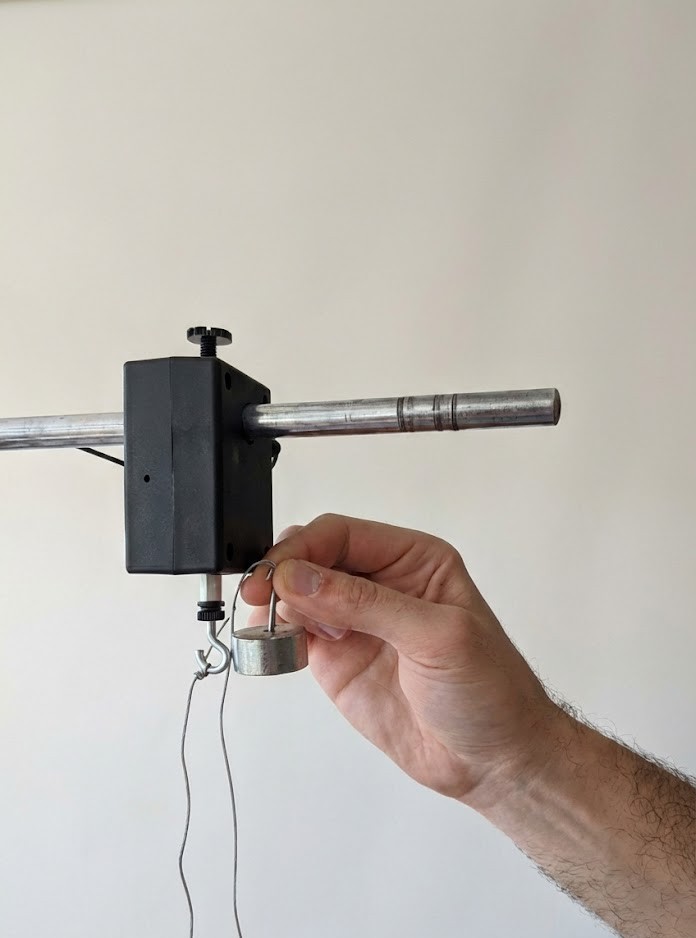 Hand hält kleines Metallgewicht an Haken unter einer Metallstange mit schwarzer Klemme