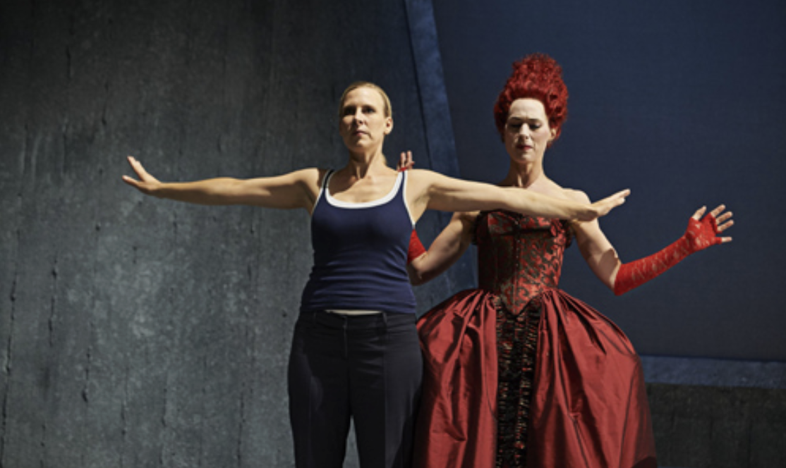 Zwei Frauen auf Bühne, eine im Tanktop, eine im roten Barockkleid, beide mit ausgestreckten Armen