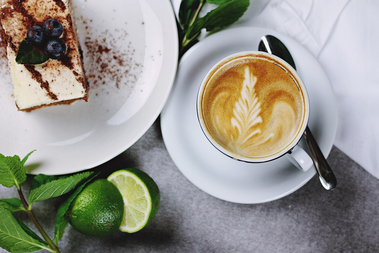 Eine Tasse Kaffee mit Latte Art, ein Stück Kuchen und Limetten auf einem Tisch.