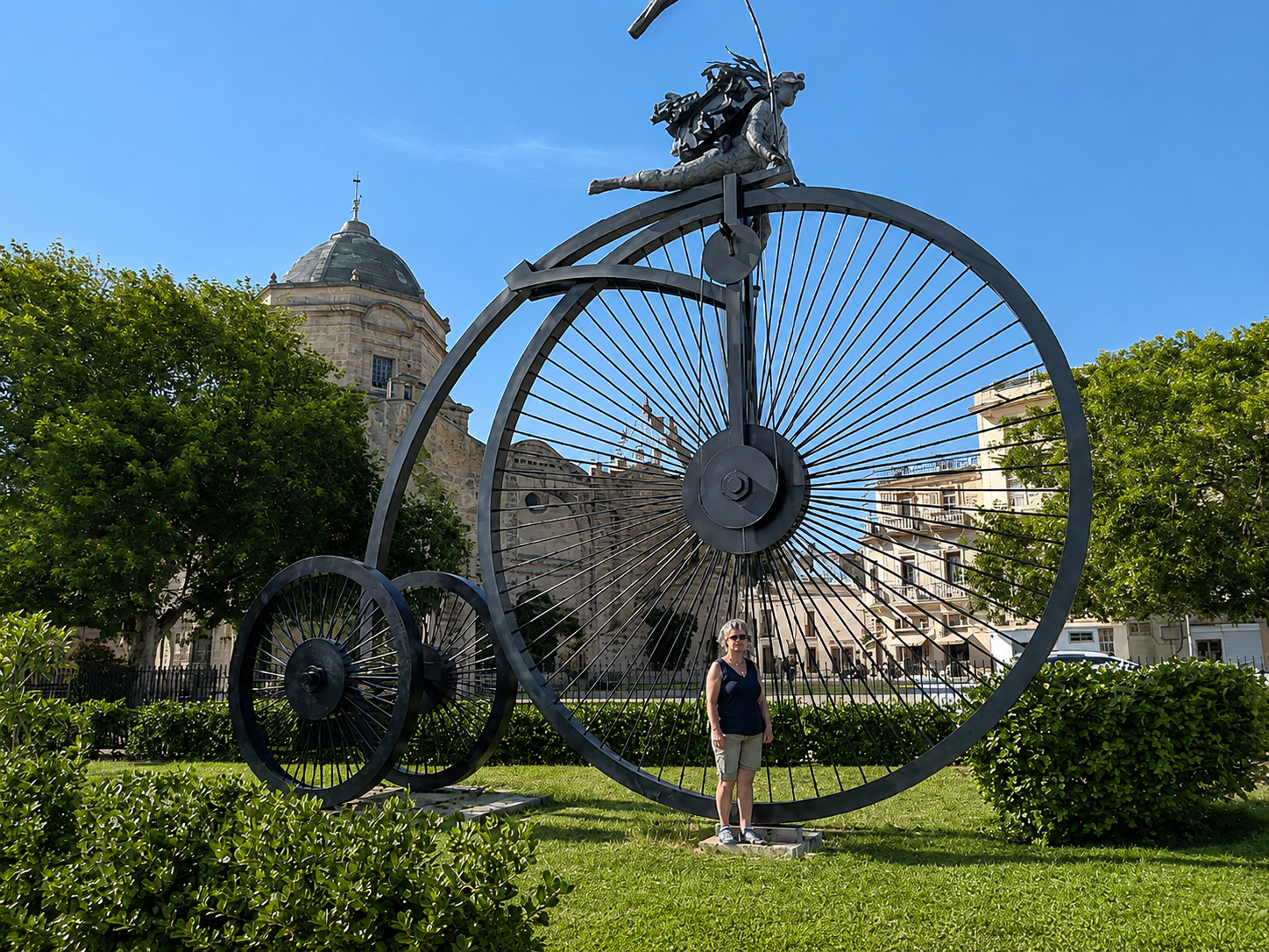 Große Metallskulptur eines Hochrads im Park, eine Person steht davor, Bäume und ein Kirchengebäude im Hintergrund.