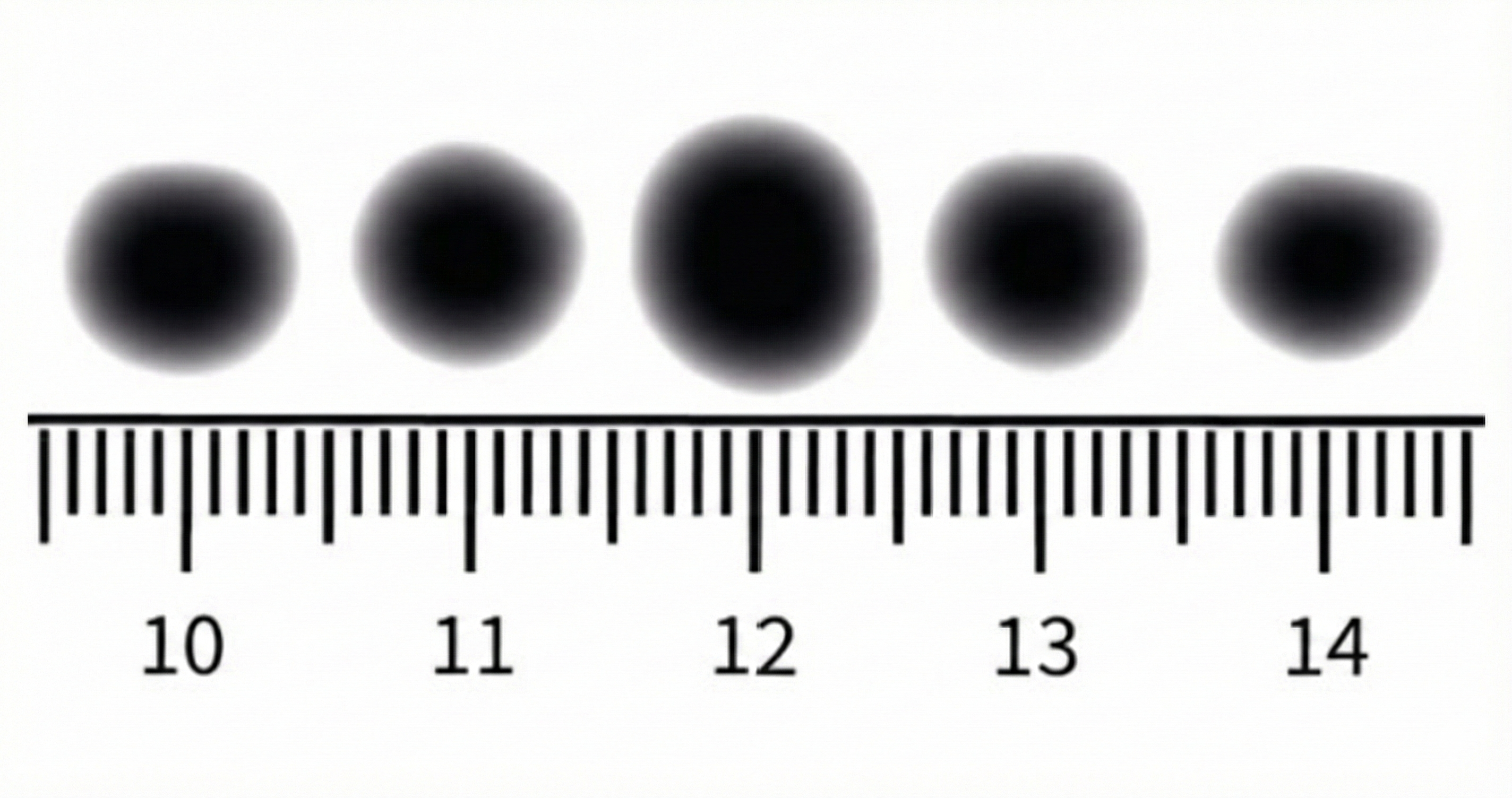 Fünf unscharfe schwarze Punkte über einem Lineal (Skala 10–14) als Größenvergleich