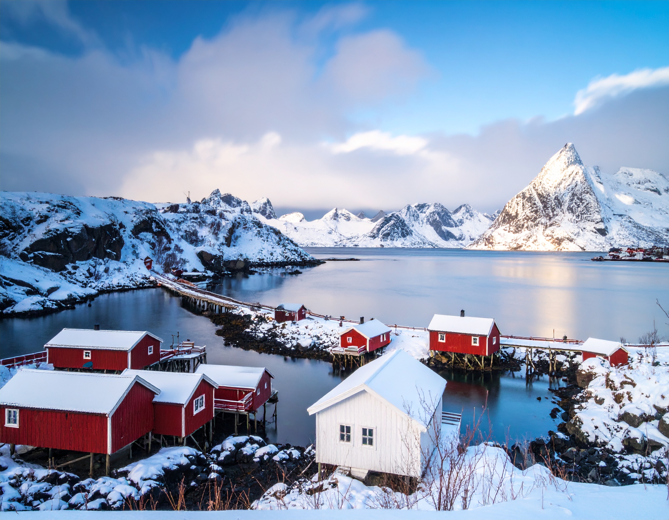 Schneebedeckte Landschaft mit roten Holzhäusern und Bergen im Hintergrund, reflektierend im ruhigen Wasser.