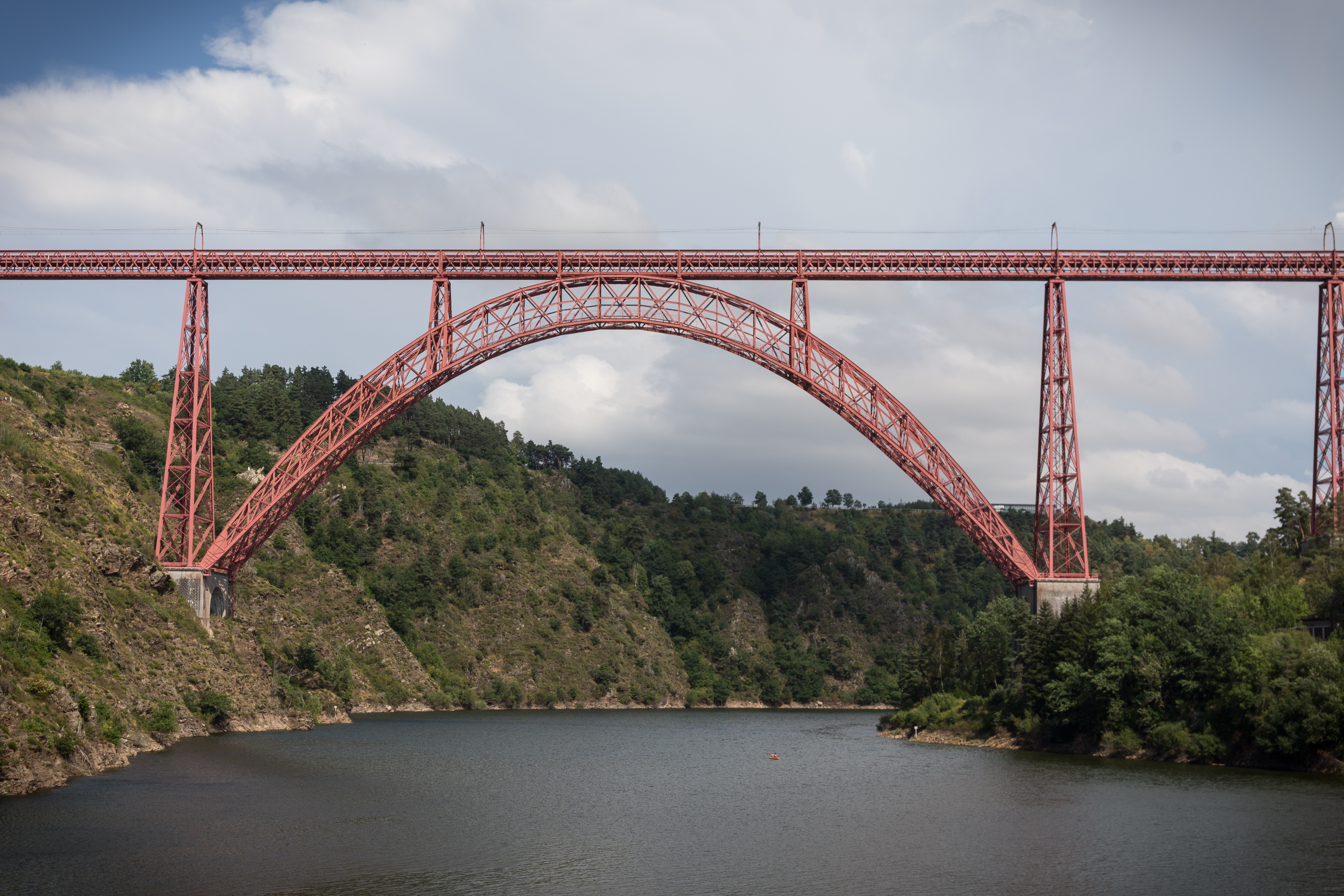 Rote Stahlbogenbrücke über ruhigem Fluss zwischen bewaldeten Hängen unter leicht bewölktem Himmel
