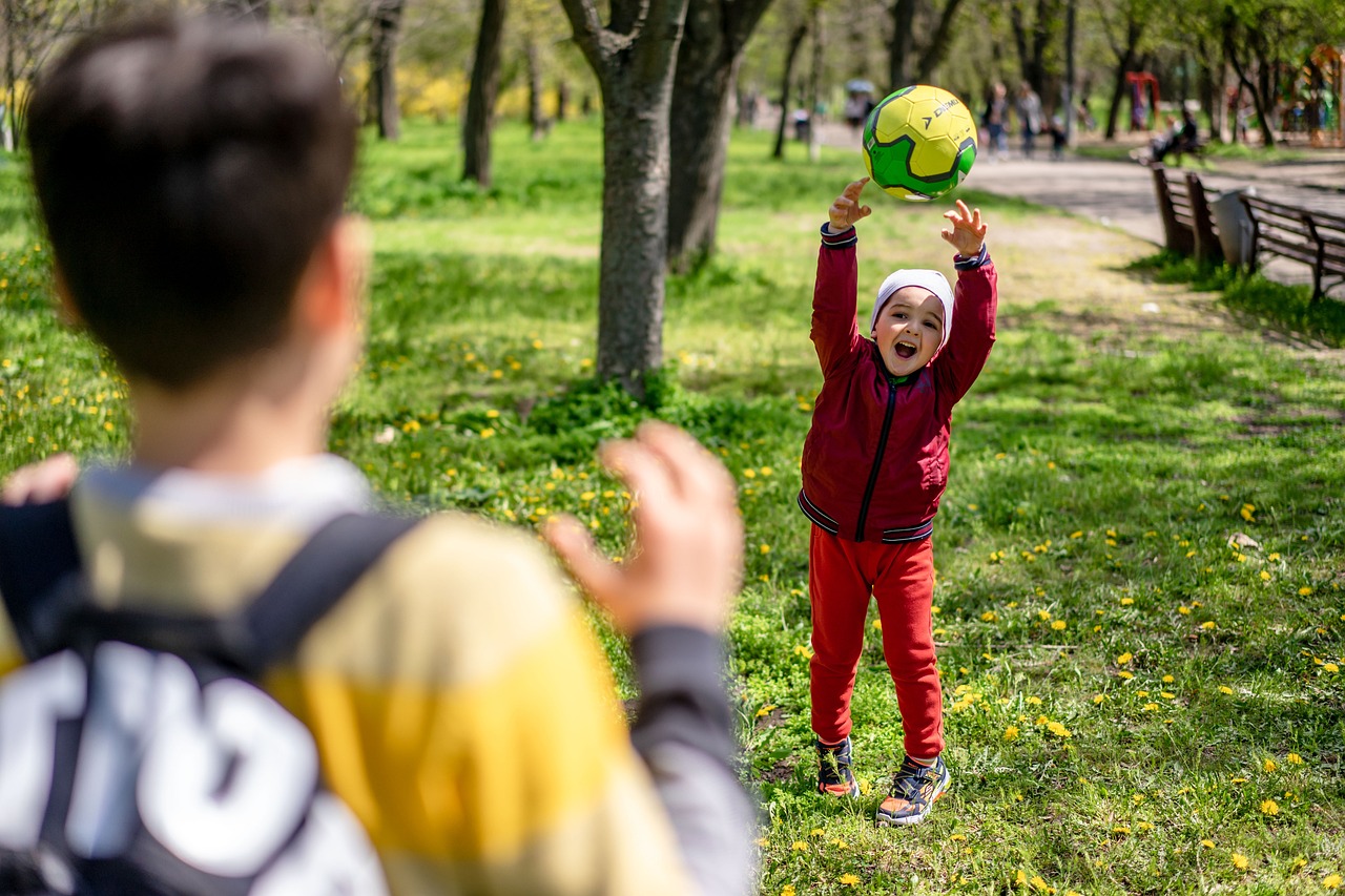 Ein Kind wirft einen Ball in einem Park und lächelt, während ein anderer Junge im Hintergrund zuschaut.