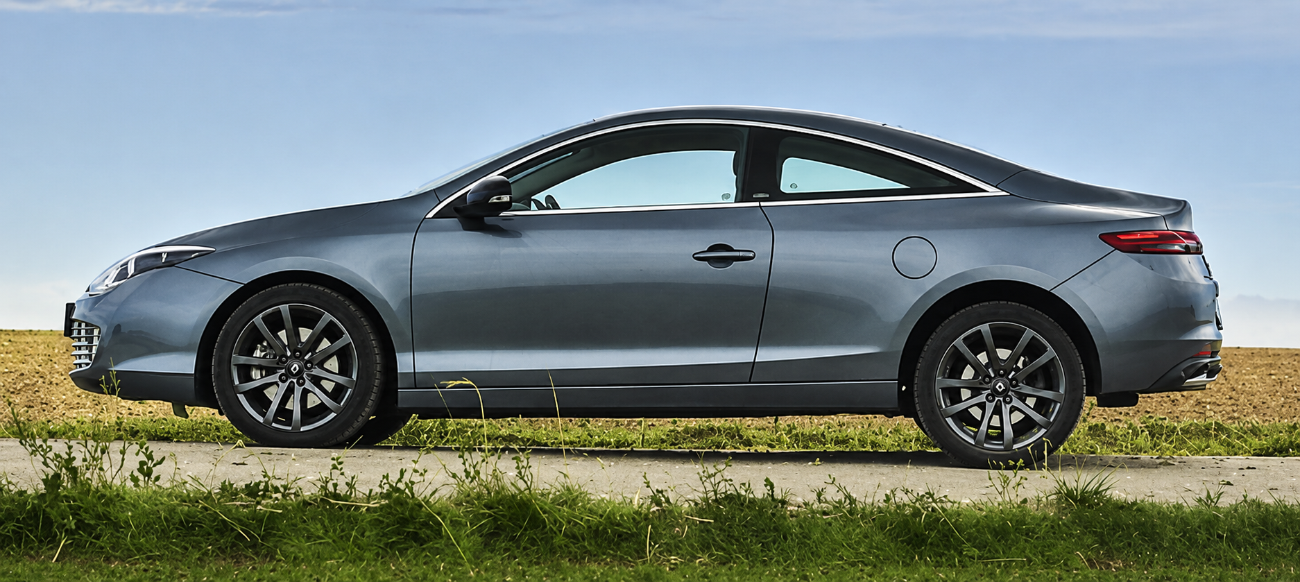 Graues Coupé seitlich auf Landstraße, Gras im Vordergrund und blauer Himmel.
