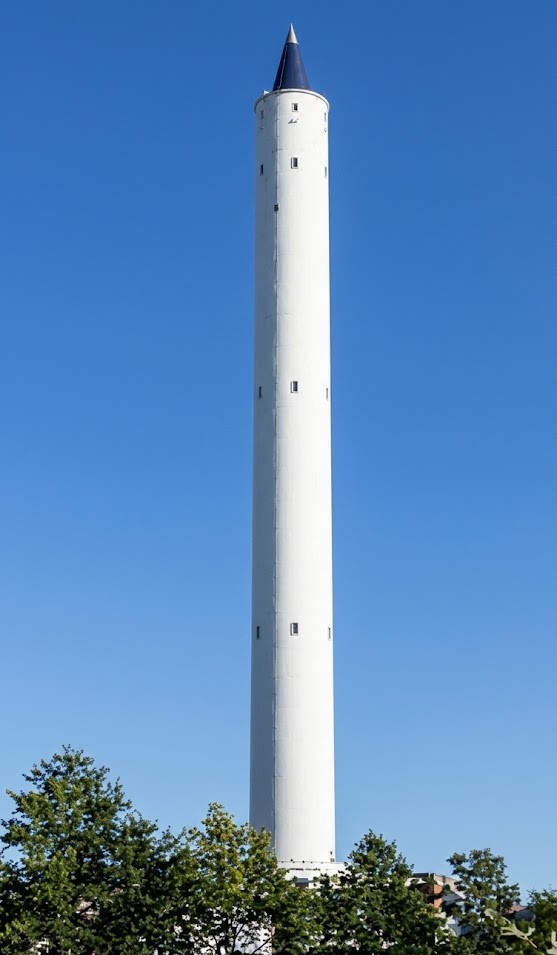 Hoher weißer Turm mit blauer Spitze vor klarem blauem Himmel, Bäume im Vordergrund.
