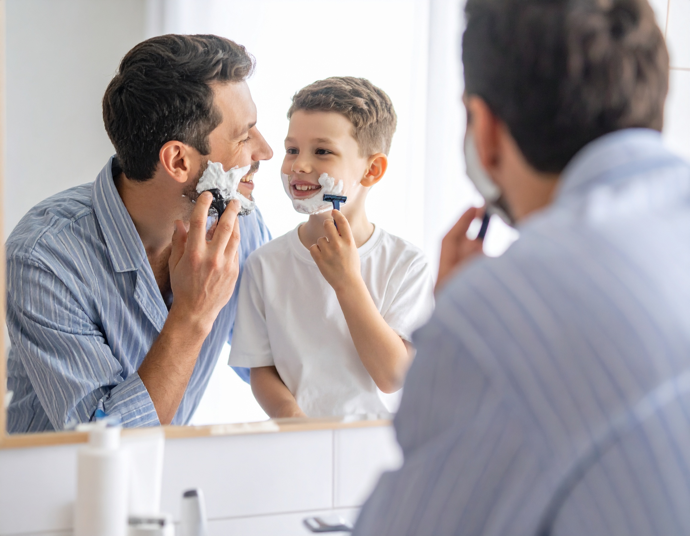 Vater und Sohn beim Rasieren im Badezimmer, beide mit Rasierschaum im Gesicht und lächelnd.