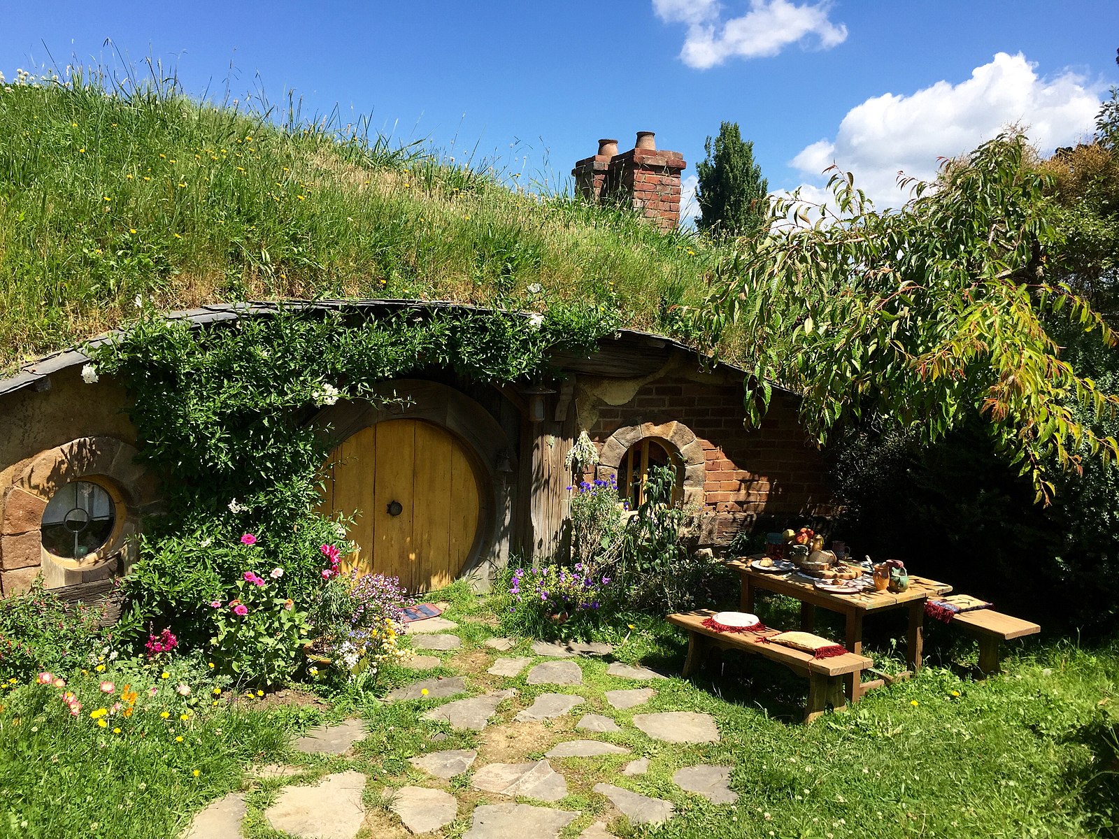 Idyllisches Hobbit-Haus mit Blumen, Grasdach und einem einladenden Tisch im Freien unter blauem Himmel.