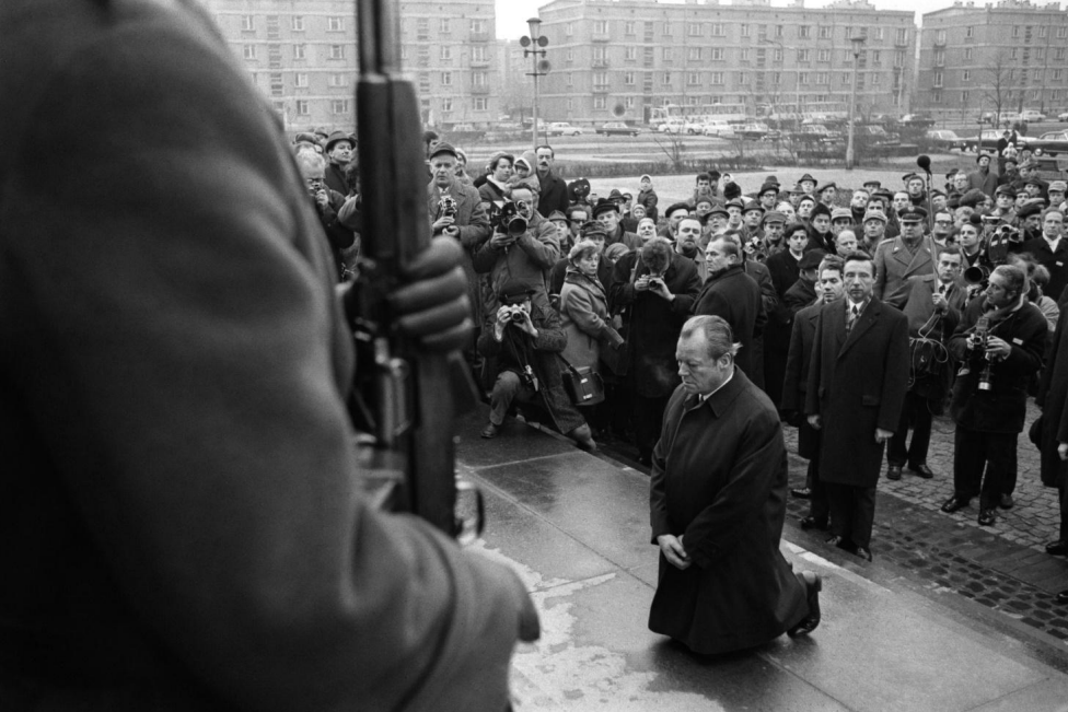 Mann kniet vor Mahnmal, bewaffneter Soldat im Vordergrund, Menschenmenge und Fotografen im Hintergrund