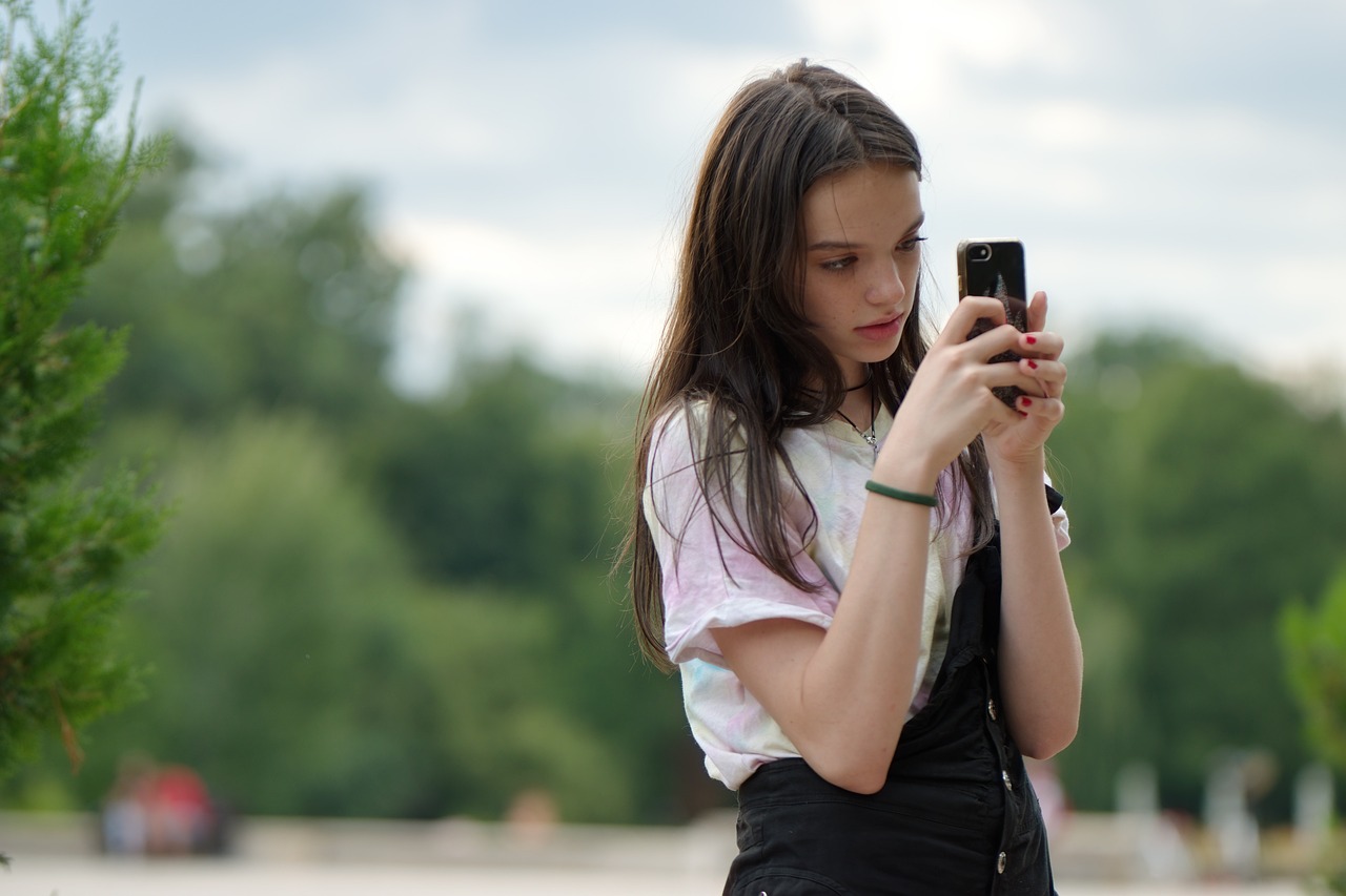 Ein Mädchen mit langen Haaren schaut auf ihr Handy, umgeben von Bäumen und einer bewölkten Himmel.