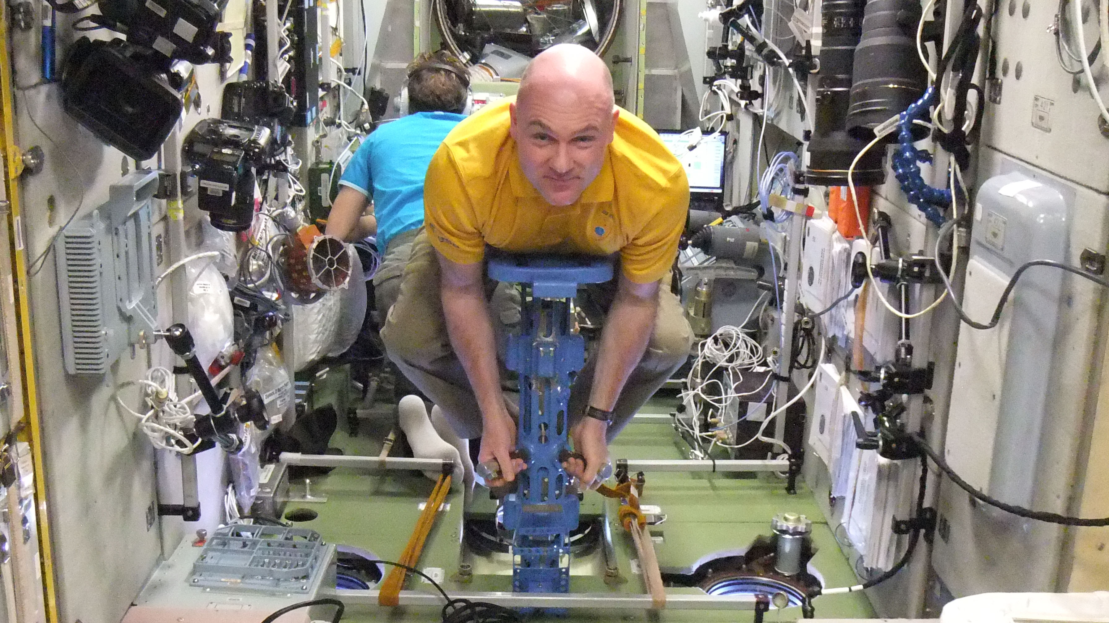 Astronaut in Raumstation auf blauem Sitzgerät zwischen Kabeln und technischen Geräten