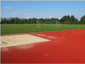 Sportplatz mit rotem Laufbahn, Sandgrube und Fußballtoren im Hintergrund.